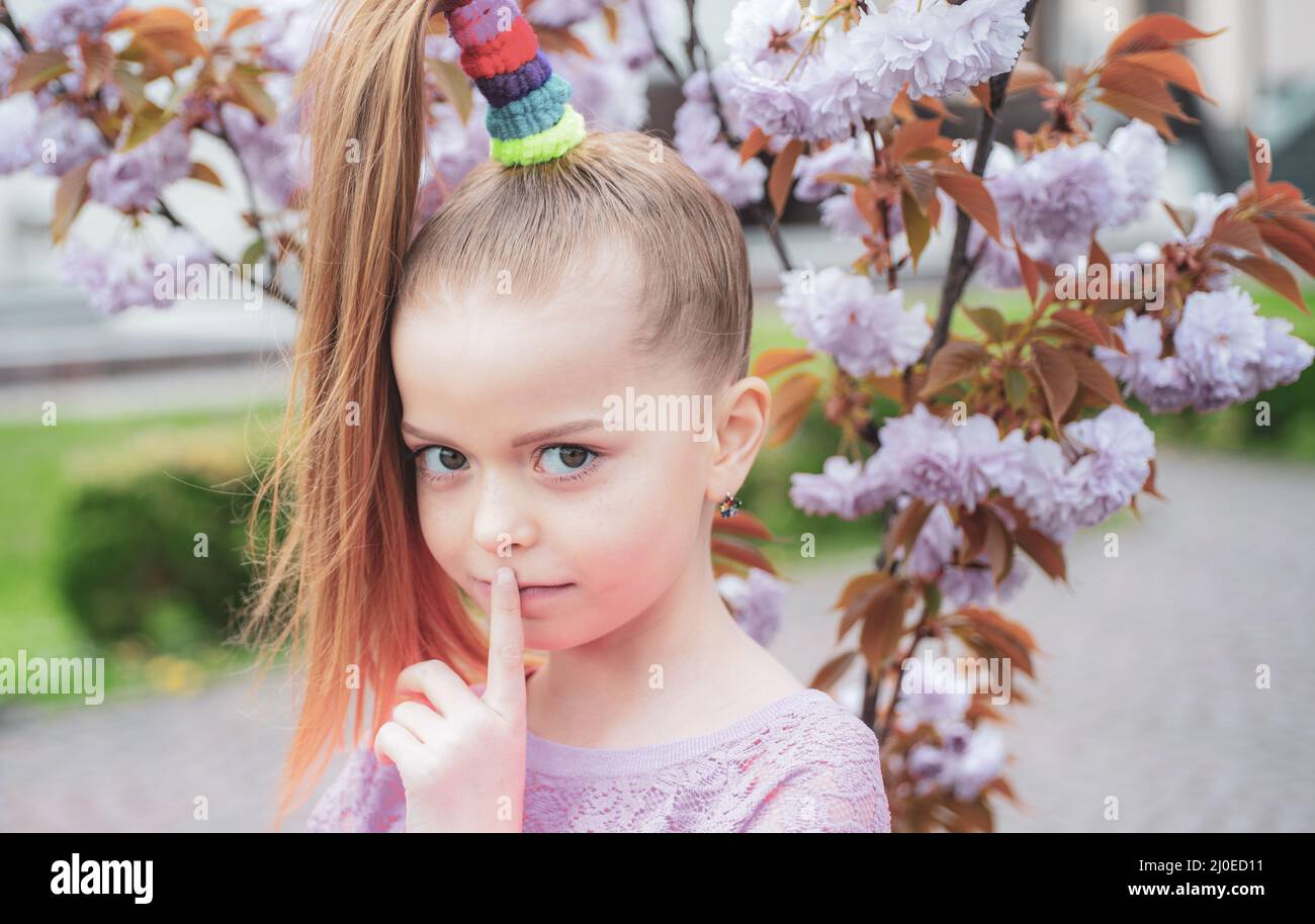 Positive schoolgirl with a long haircut holding her finger at the lips. Child secrets. Blooming sakura cherry. Stock Photo
