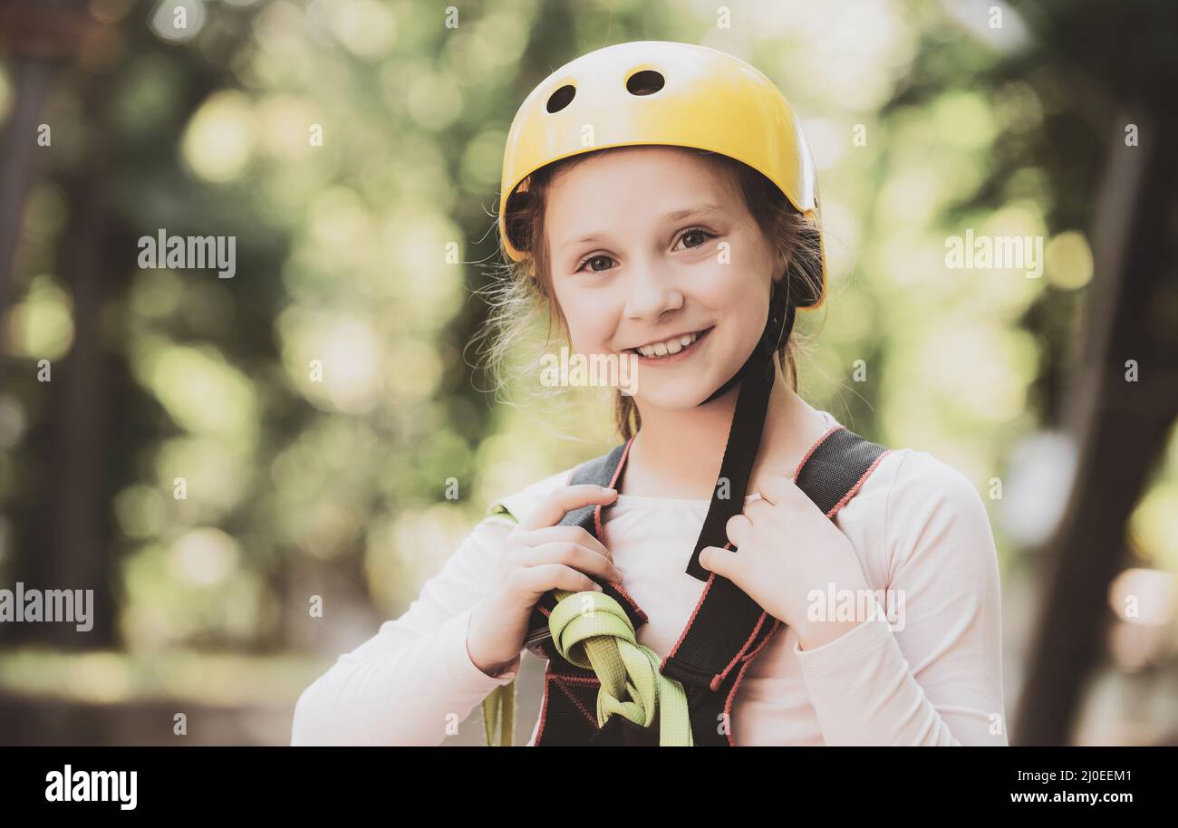 Portrait of a beautiful kid on a rope park among trees. Happy Little girl climbing a tree. Helmet - safety equipment for Little girl playing Stock Photo