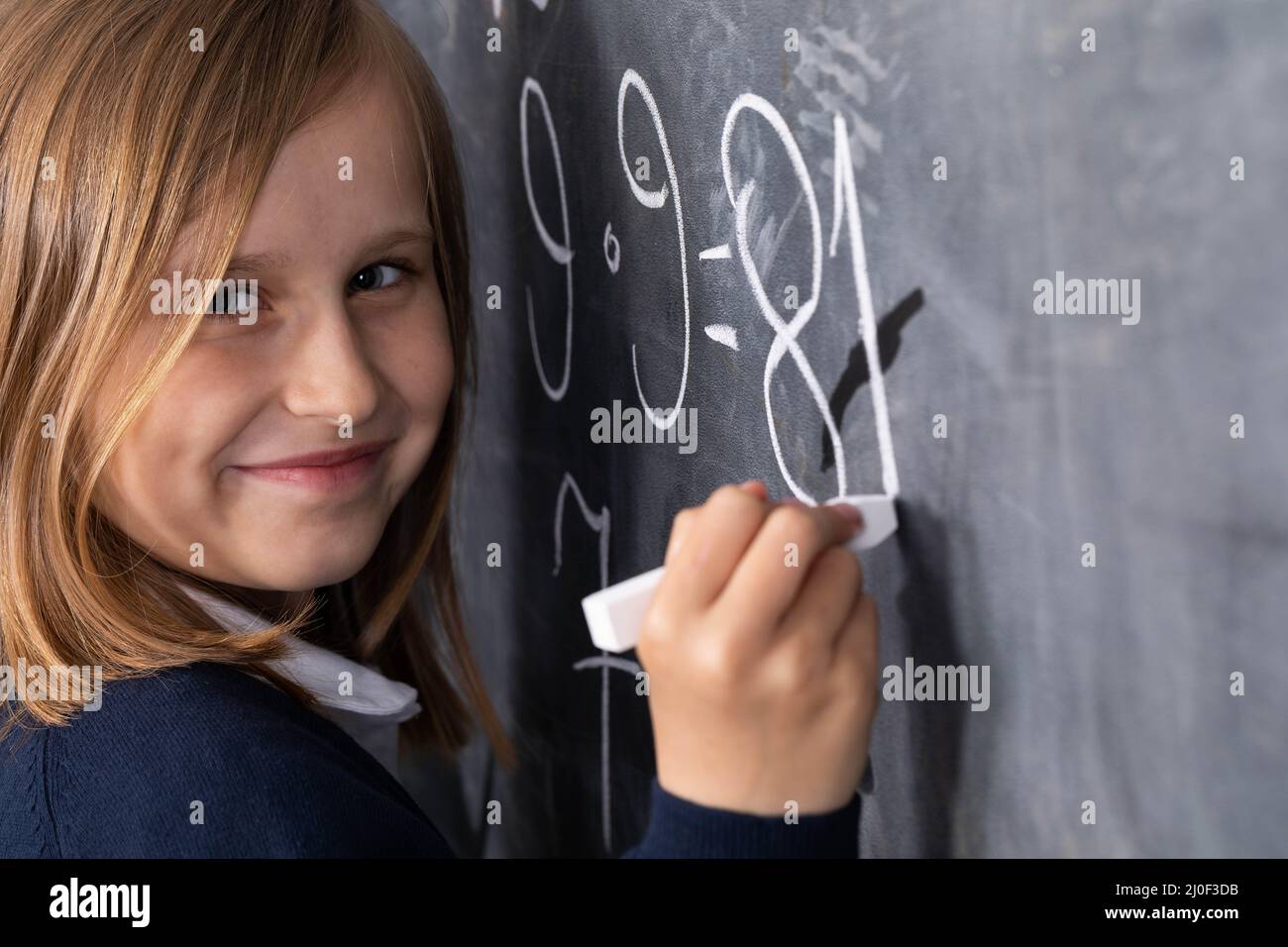 The student solves a math problem standing at the blackboard in her classroom. Stock Photo