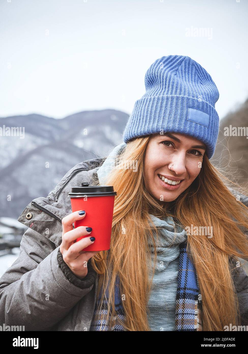 Smiling red head girl in blue hat holding red paper cup with coffee on blurred landscape background Stock Photo