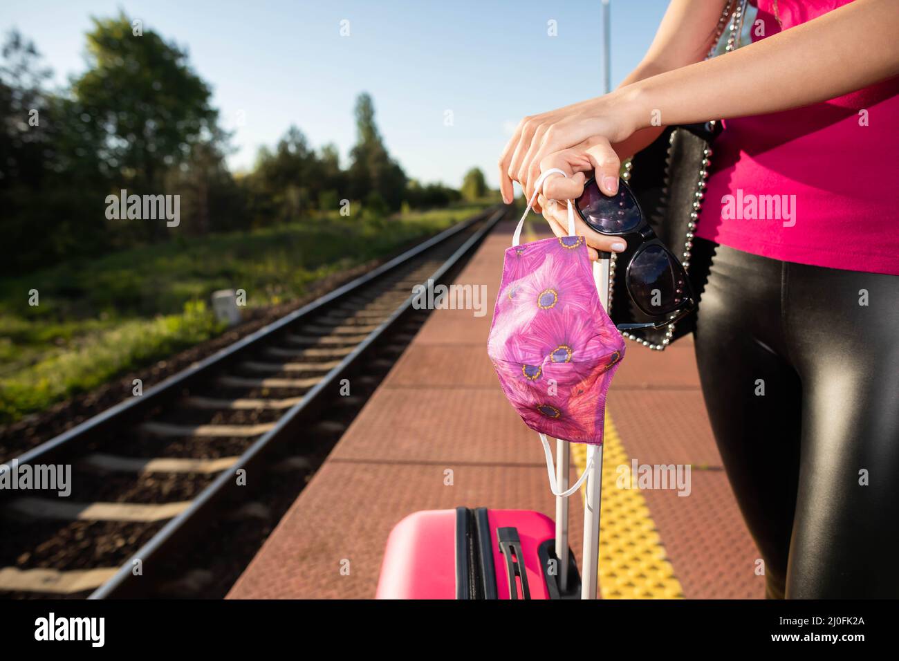 A teenage girl is standing in a face mask on a train platform waiting for her train. A summer adventure in rail travel. Stock Photo