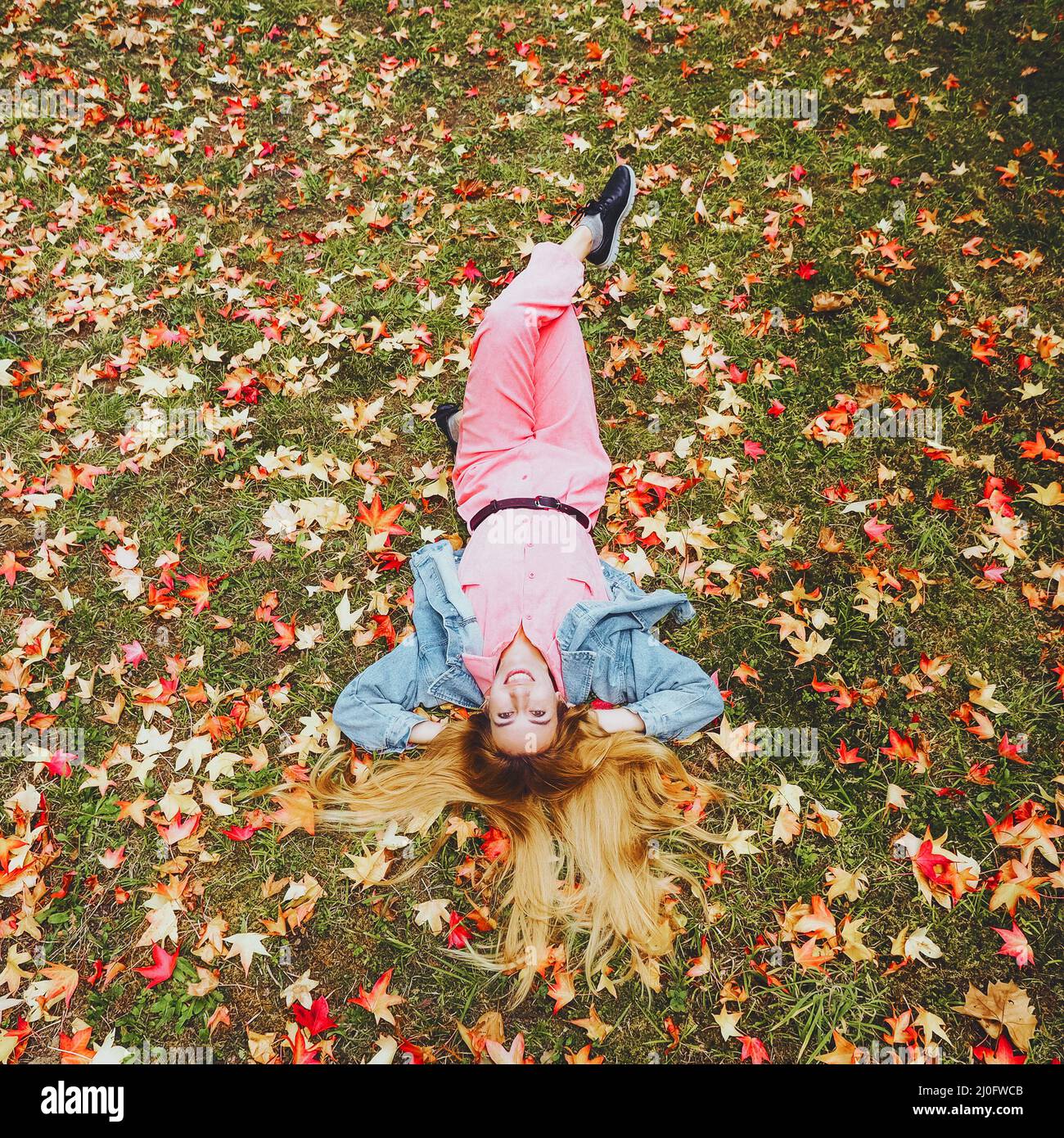 A red-haired girl in a pink jumpsuit lies with her hair outstretched in a meadow covered with autumn maple leaves Stock Photo