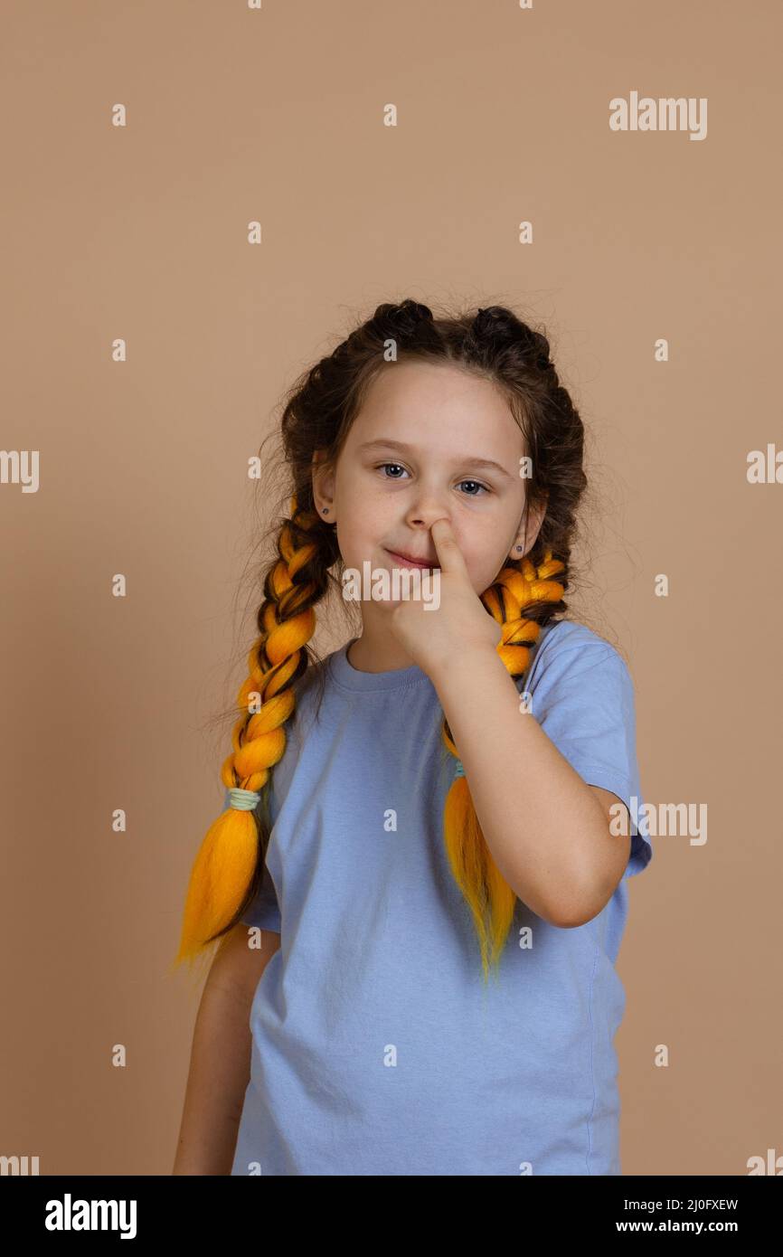 Caucasian mischievous young happy girl looking at camera with slight smile picking nose with finger having kanekalon braids on beige background Stock Photo