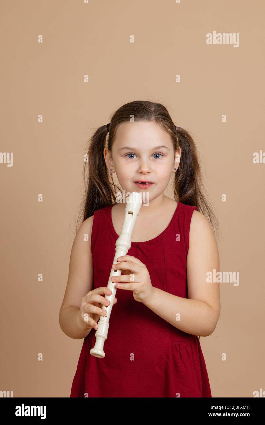 Girl in red dress thoughtfully hold common flute and pinch holes on it, beige background. Learning to play woodwind musical instrument. Stock Photo