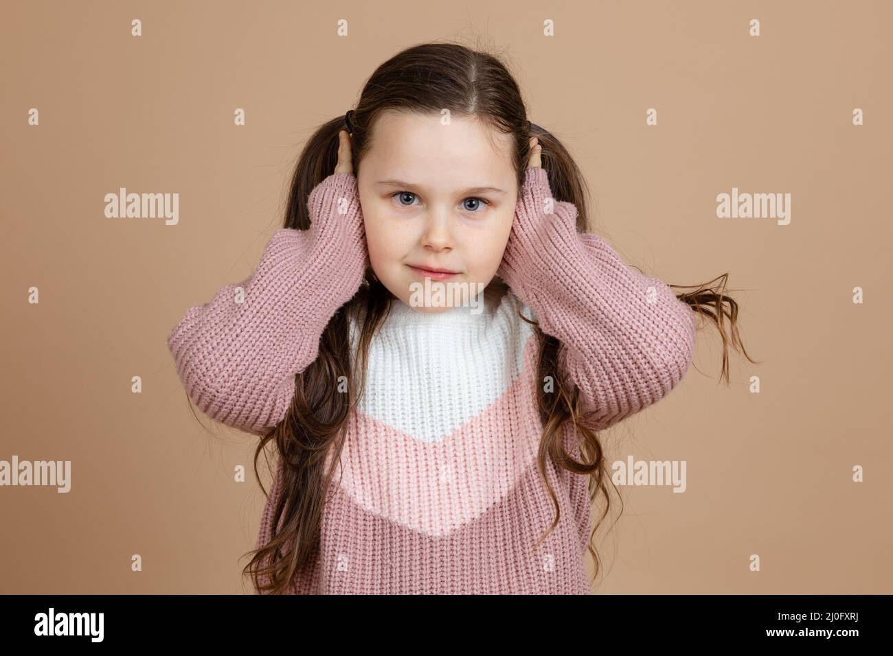 Portrait of young adorable cute smiling girl with long dark hair in white, pink sweater standing, covering ears with hands, posing. Stock Photo