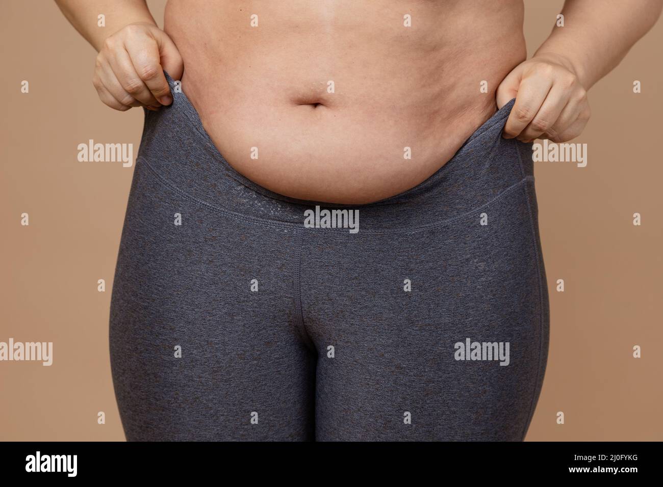 Closeup of woman putting on leggings with flabby large abdomen on beige background. Body positive. Violation of cell elasticity, loss of nutrients Stock Photo