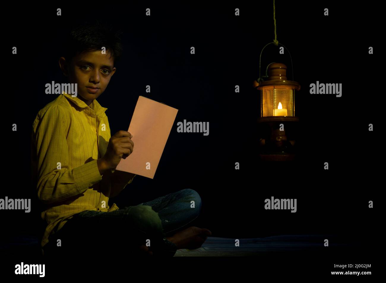 high ISO shot, young school kid looking at camera with book in hand under oil lamp or lantern due to power loss - concpet of poverty, blackout during Stock Photo
