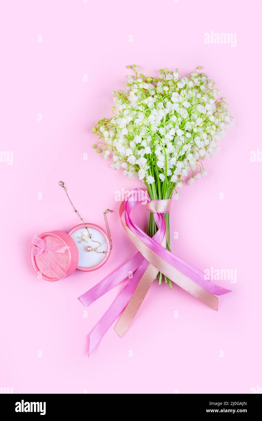 A bouquet of lily of the valley flowers with pink ribbons and a pink box with a gold bracelet on a pink background close up Stock Photo