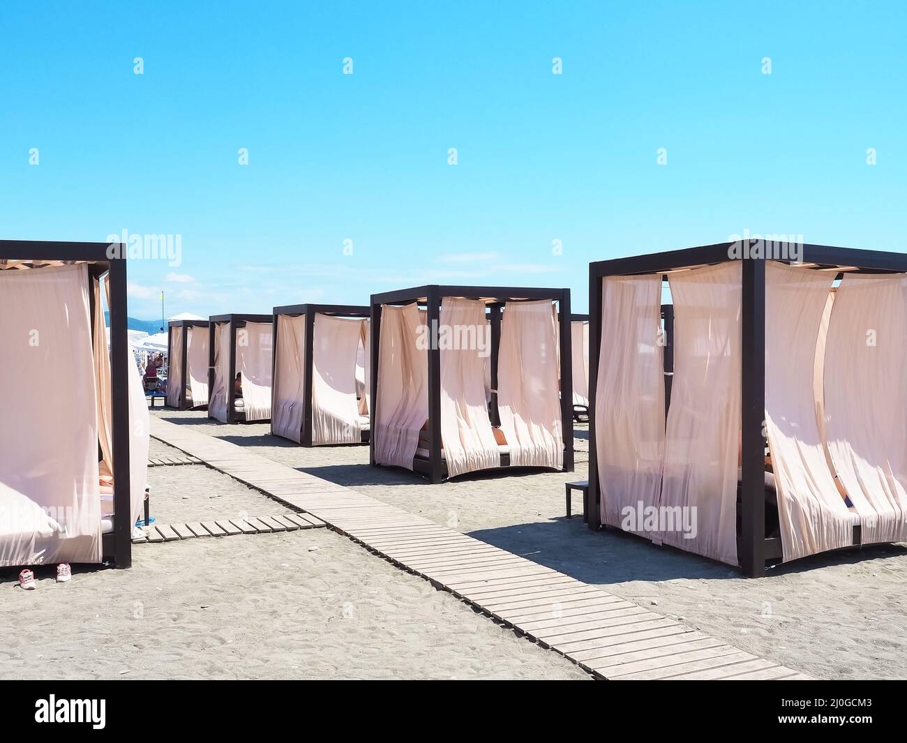 Beach cabins with a metal frame and light curtains stand in a row on the sand with wooden paths between them Stock Photo