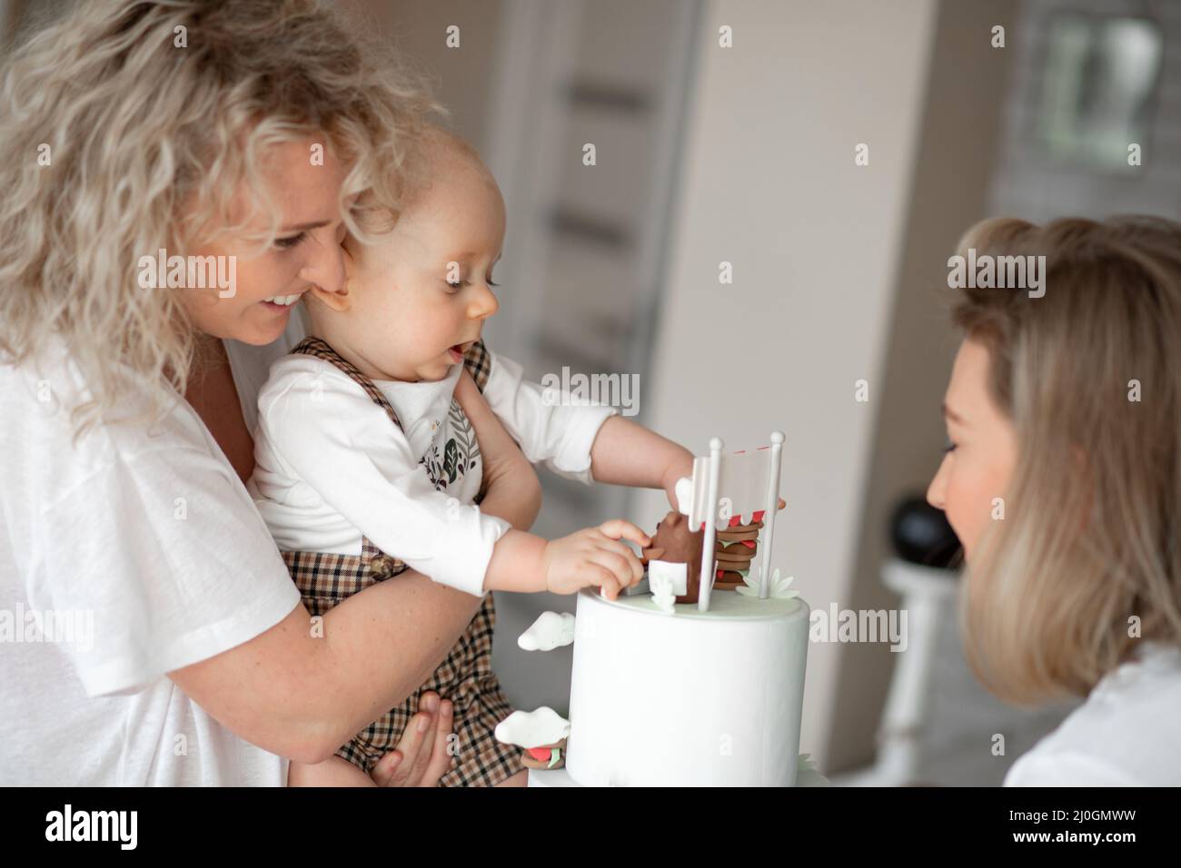 Two smiling beauteous middle-aged and young women congratulate cute little one-year-old boy touching bear on white cake. Stock Photo