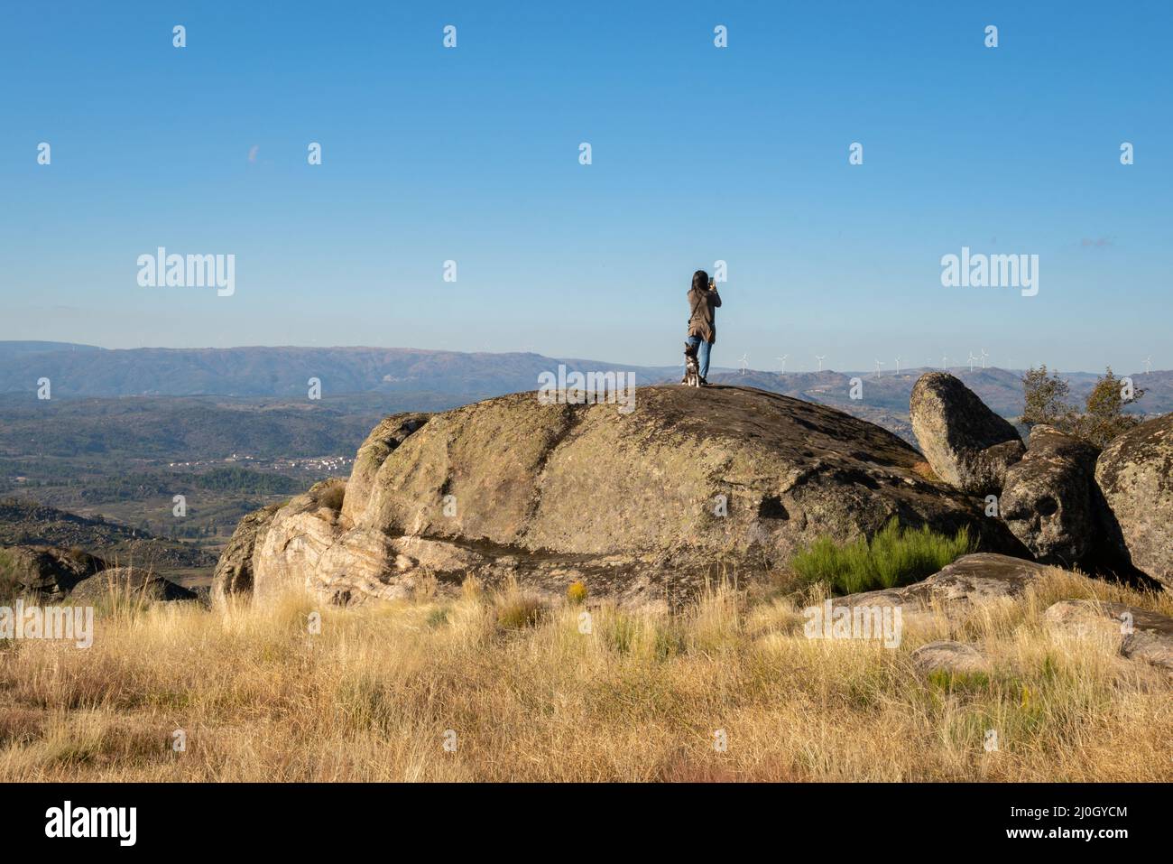 Caucasian young woman with brown dog on top of a boulder stone seeing Sortelha nature mountain landscape, in Portugal Stock Photo