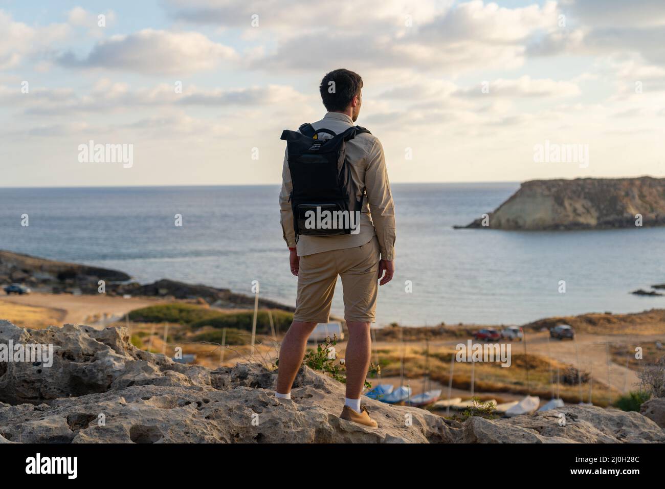 Man with backpack is hiking along a rocky coast at sunset near the Mediterranean sea in Cyprus. Male tourist admires the view Ye Stock Photo