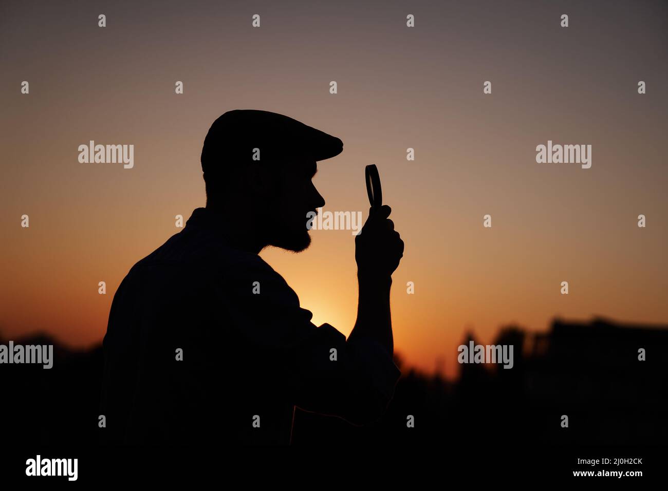 Silhouette portrait of man investigator in peaking cap looking with magnifying at side. Man searching sales or discounts at sunset using loupe with sun on background. High quality image Stock Photo