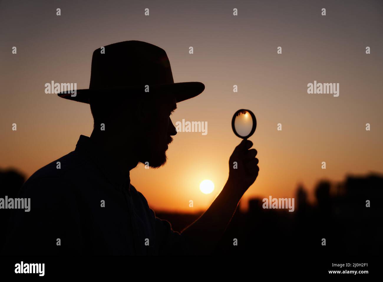 Man searching using loupe with sun on background and urban view. Male detective or investigator silhouette portrait in hat looking with magnifying at side. Investigation or inspection concept Stock Photo