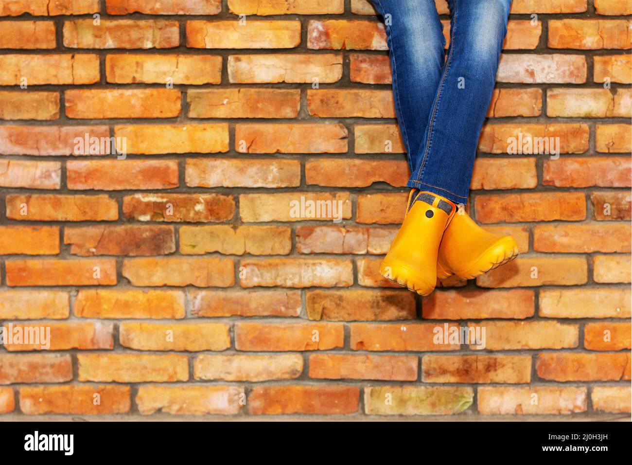 Cross-legged sitting on a brick wall of a young girl in jeans and orange galoshes against the background of a brick wall with a Stock Photo
