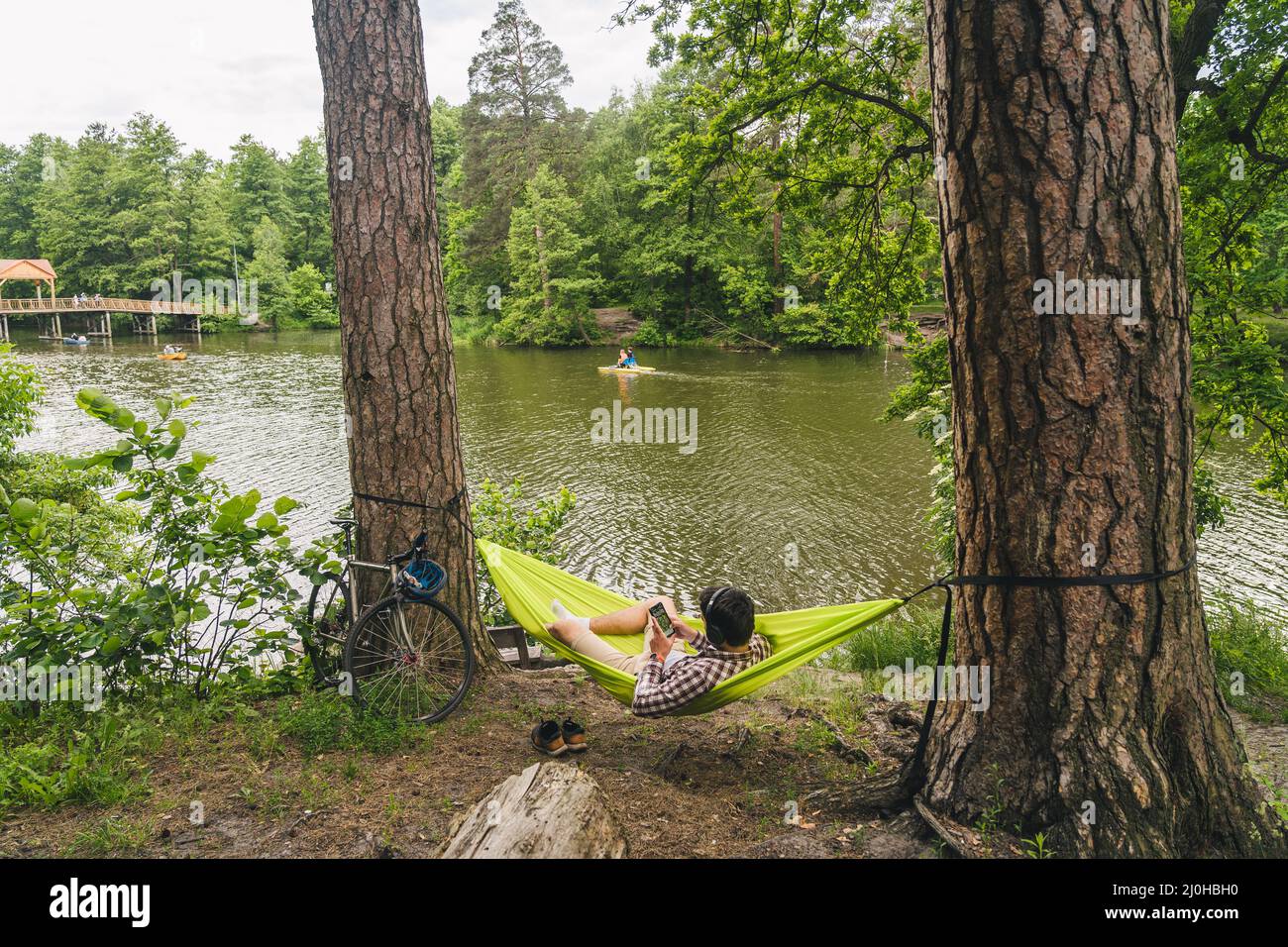 Young caucasian man resting in hammock, listening to music on headphones and using smartphone afterwards on bicycle in forest ne Stock Photo
