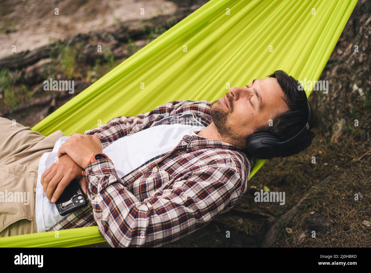 Man on bicycle trip at camping by lake is relaxing in green hammock while listening to music. Active recreation theme in nature. Stock Photo