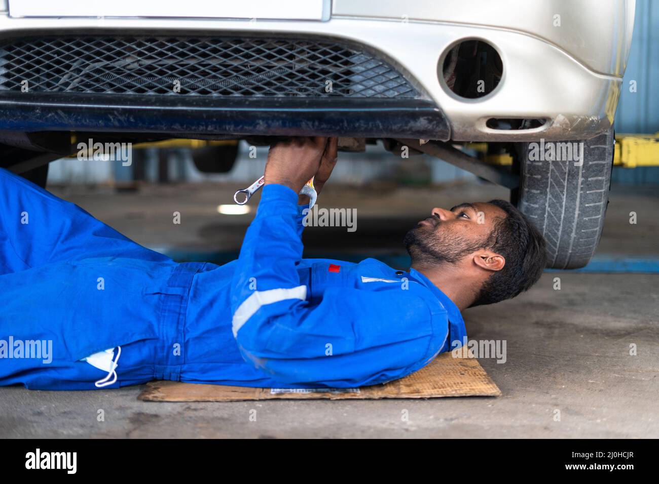 Profgessional car mechanic lying down while working under car at garage - concept of hard worker, skill labour, blue collar jobs and repair service Stock Photo