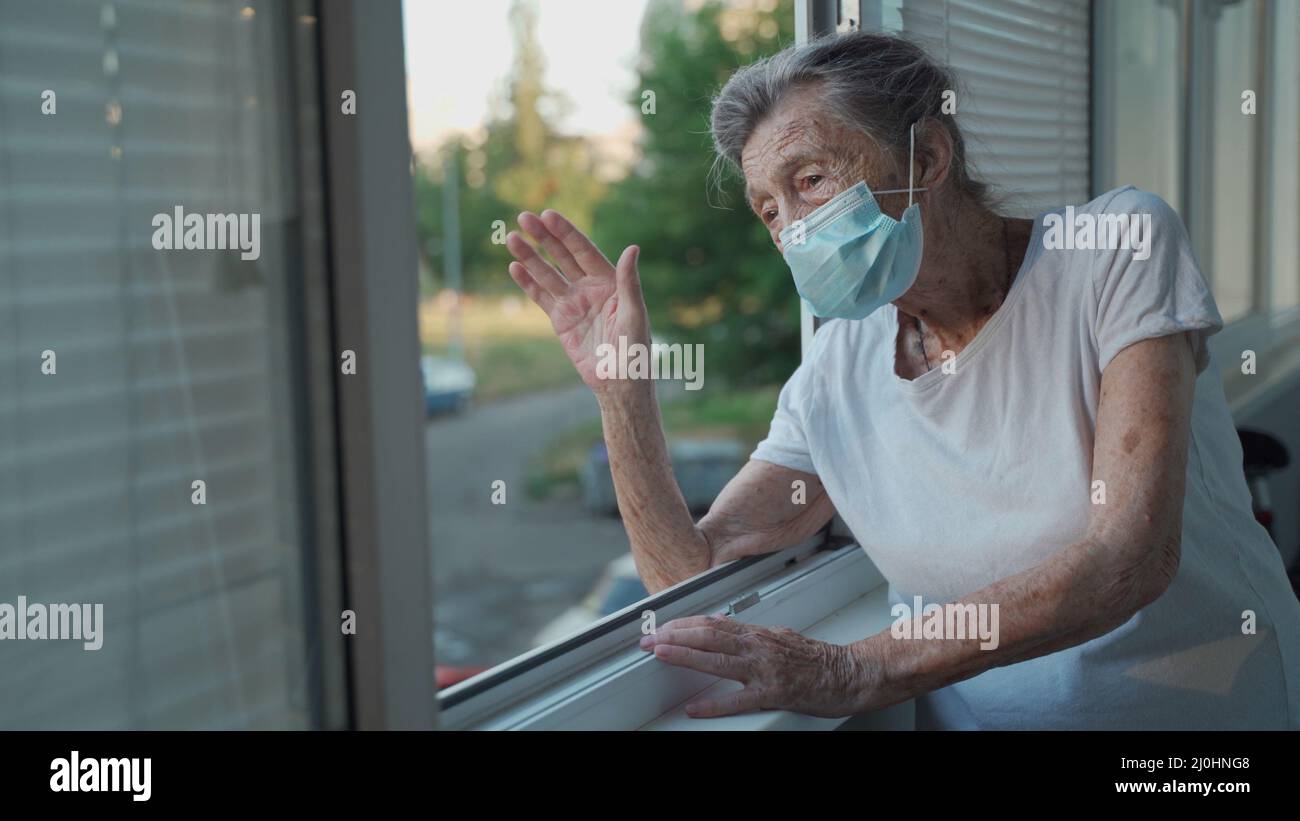 Lonely senior woman in mask stands by window and waves her hand to family who came visit her at hospital. 90 year old female in Stock Photo