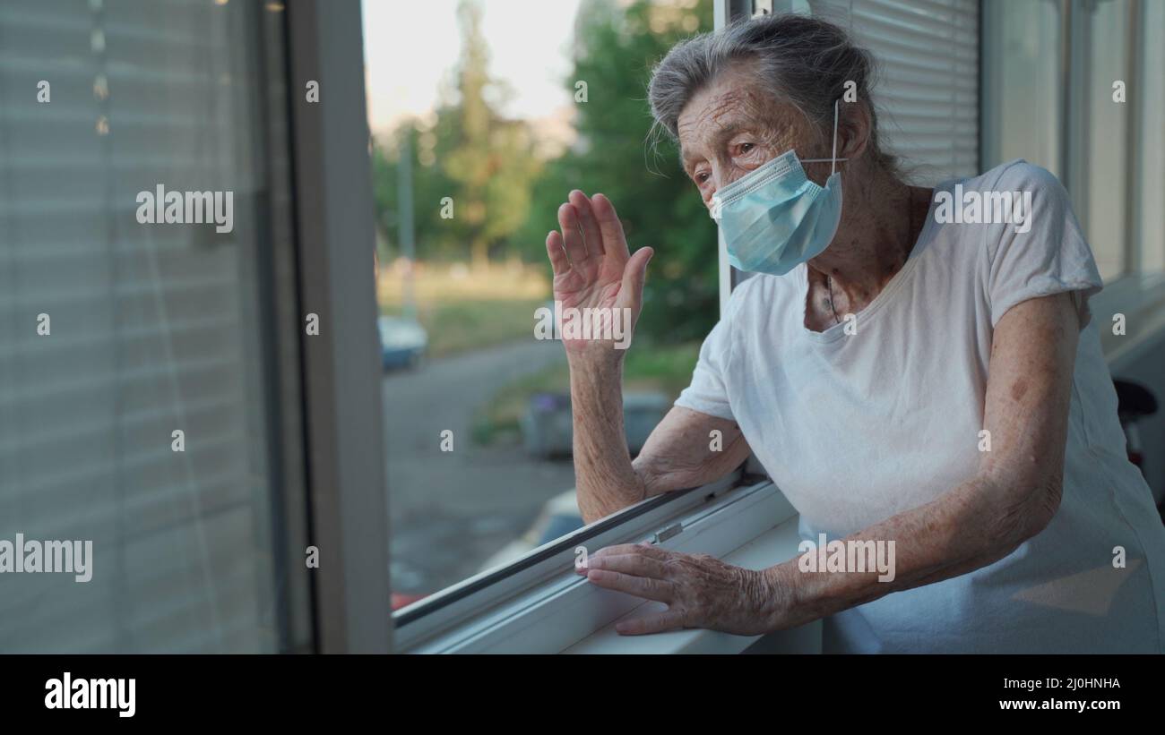 Portrait of masked senior woman in late 90s waving from home window. Elderly woman in protective mask looks out the window and w Stock Photo