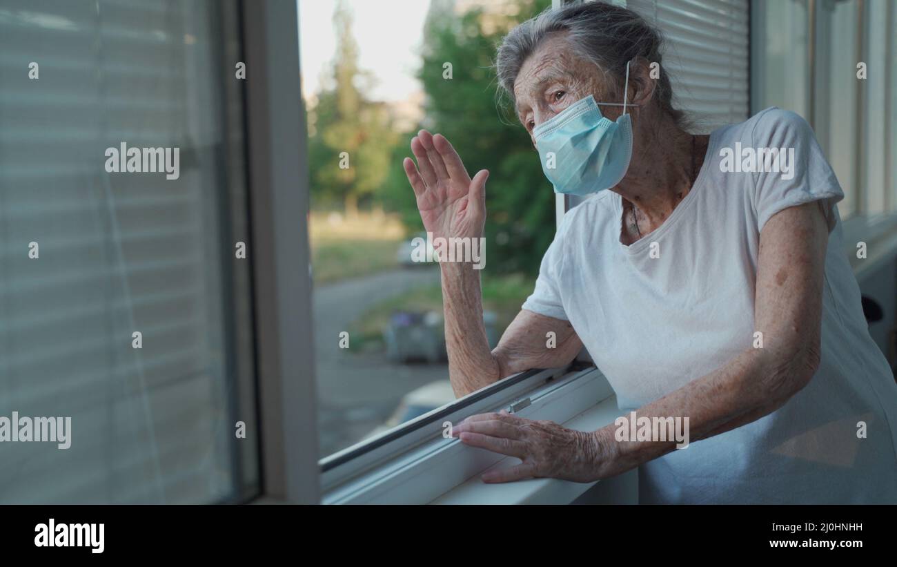 Happy senior woman in mask waves to family from window hospital. Masked elderly female glad that children came visit her in nurs Stock Photo