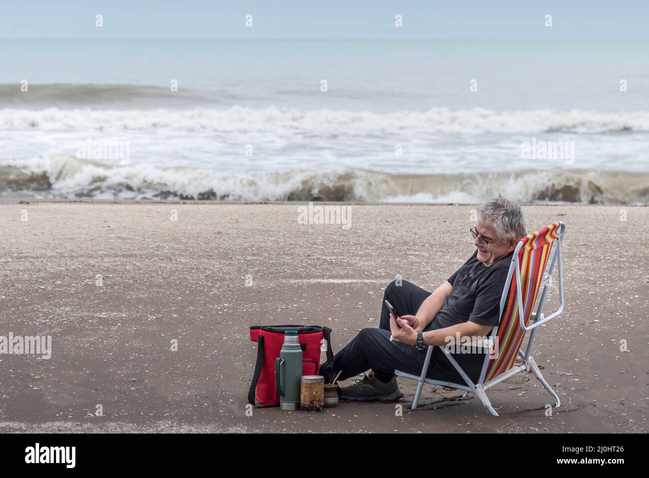 Mature man with gray hair and eyeglasses sitting on a beach chair smiling while looking at his smartphone. The waves of the sea in the background. Stock Photo