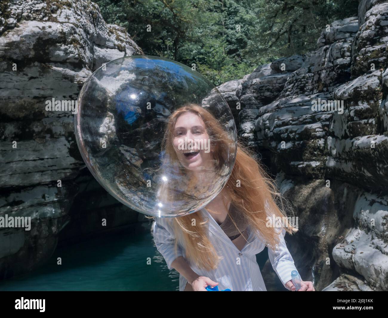 Laughing girl catches up with a shiny soap bubble looking through it against the backdrop of canyons and water Stock Photo
