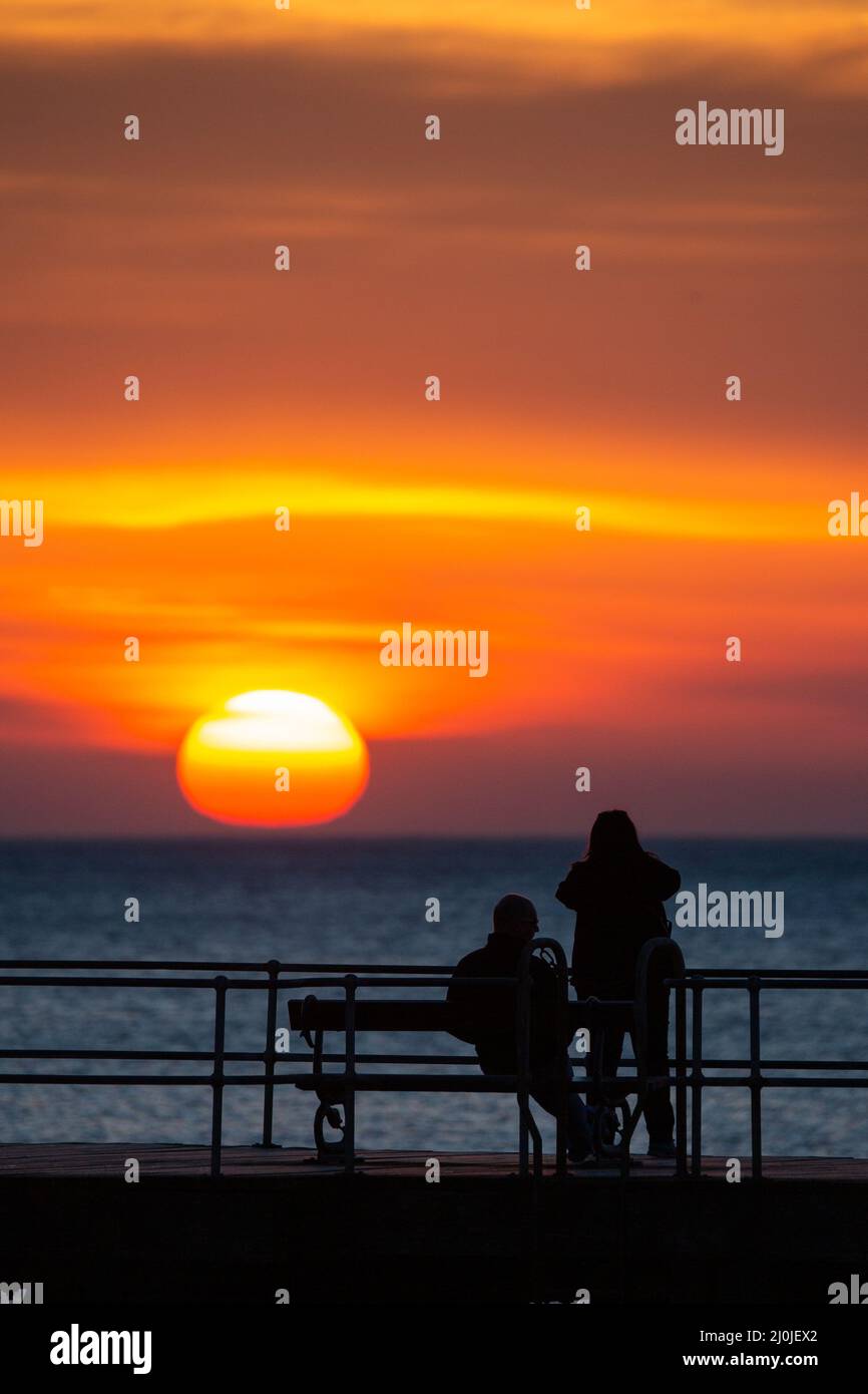 Aberystwyth, Ceredigion, Wales, UK. 19th March 2022 UK Weather: At the end of a warm day, people enjoy the view of a beautiful sunset as they walk and sit along the wooden jetty in Aberystwyth, mid Wales. © Ian Jones/Alamy Live News Stock Photo