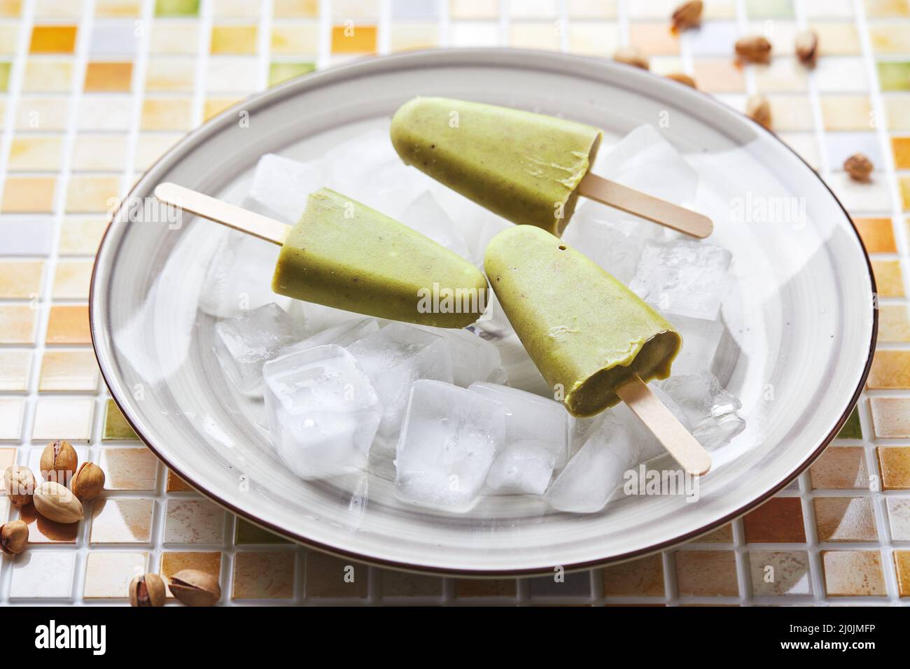 Frozen homemade pistachio popsicle in bowl of ice on mosaic tile table. Refreshing popsicle, frozen green juice on stick. Top vi Stock Photo