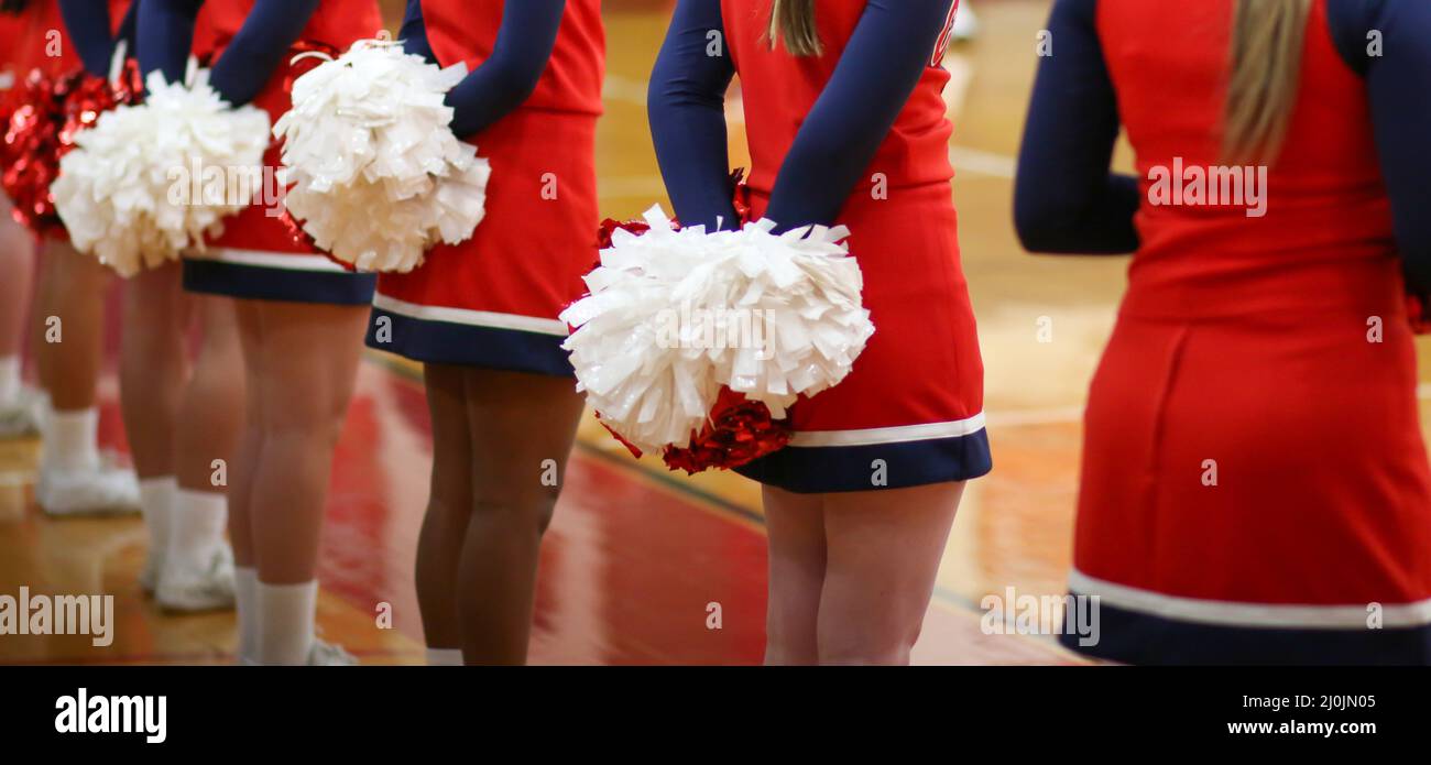 Horizontal picture of cheerleaders with their pom poms behind their backs while they watch their teams basketball game ready to cheer. Stock Photo