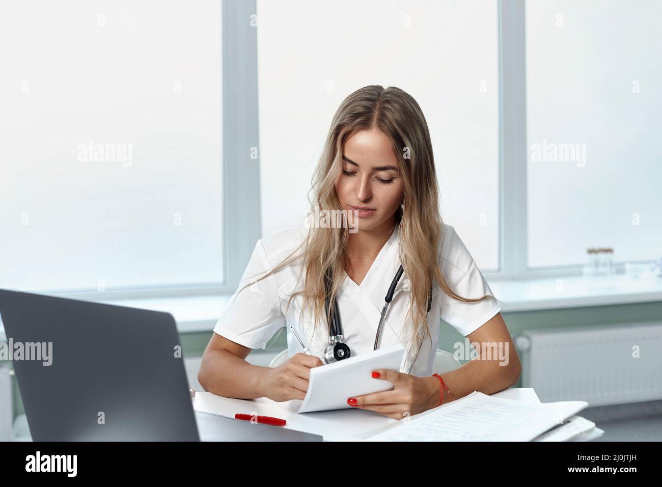 female doctor using laptop and writing notes in medical journal sitting at desk. Stock Photo