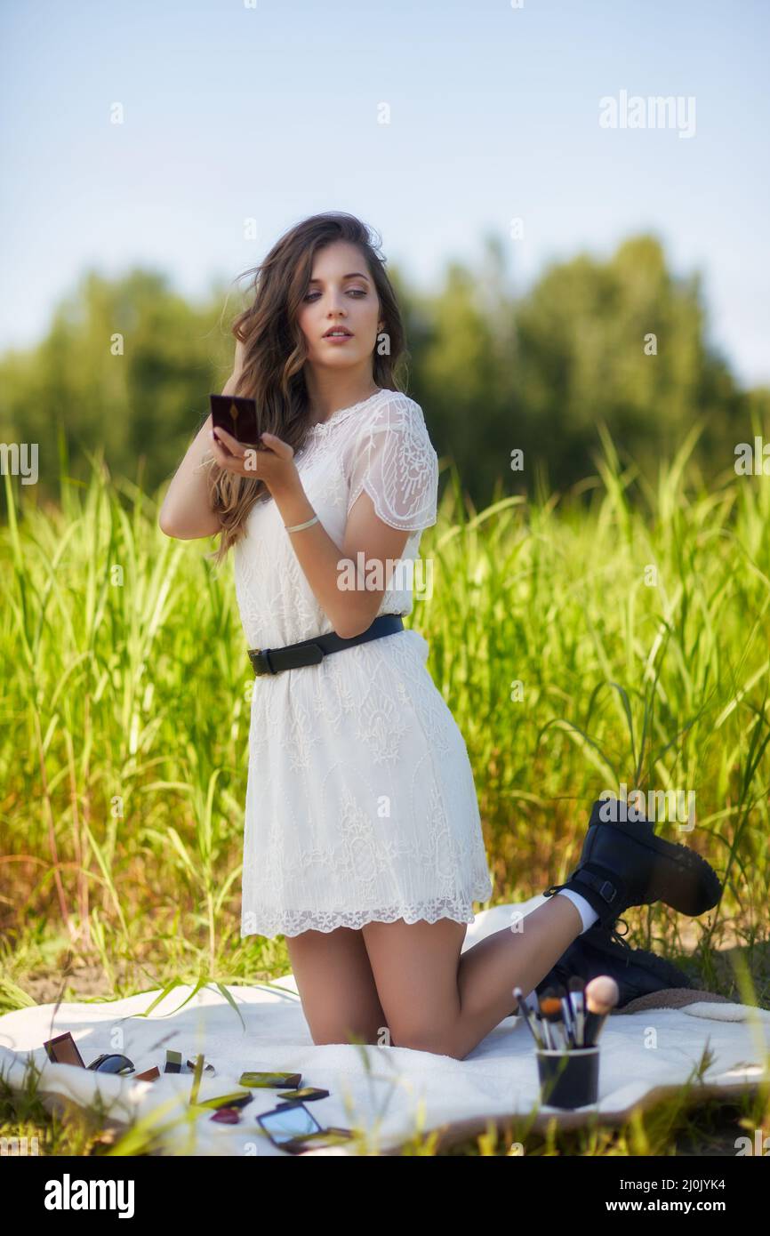 Young blonde woman in white dress is kneeling on a picnic sheet in tall grass. Stock Photo