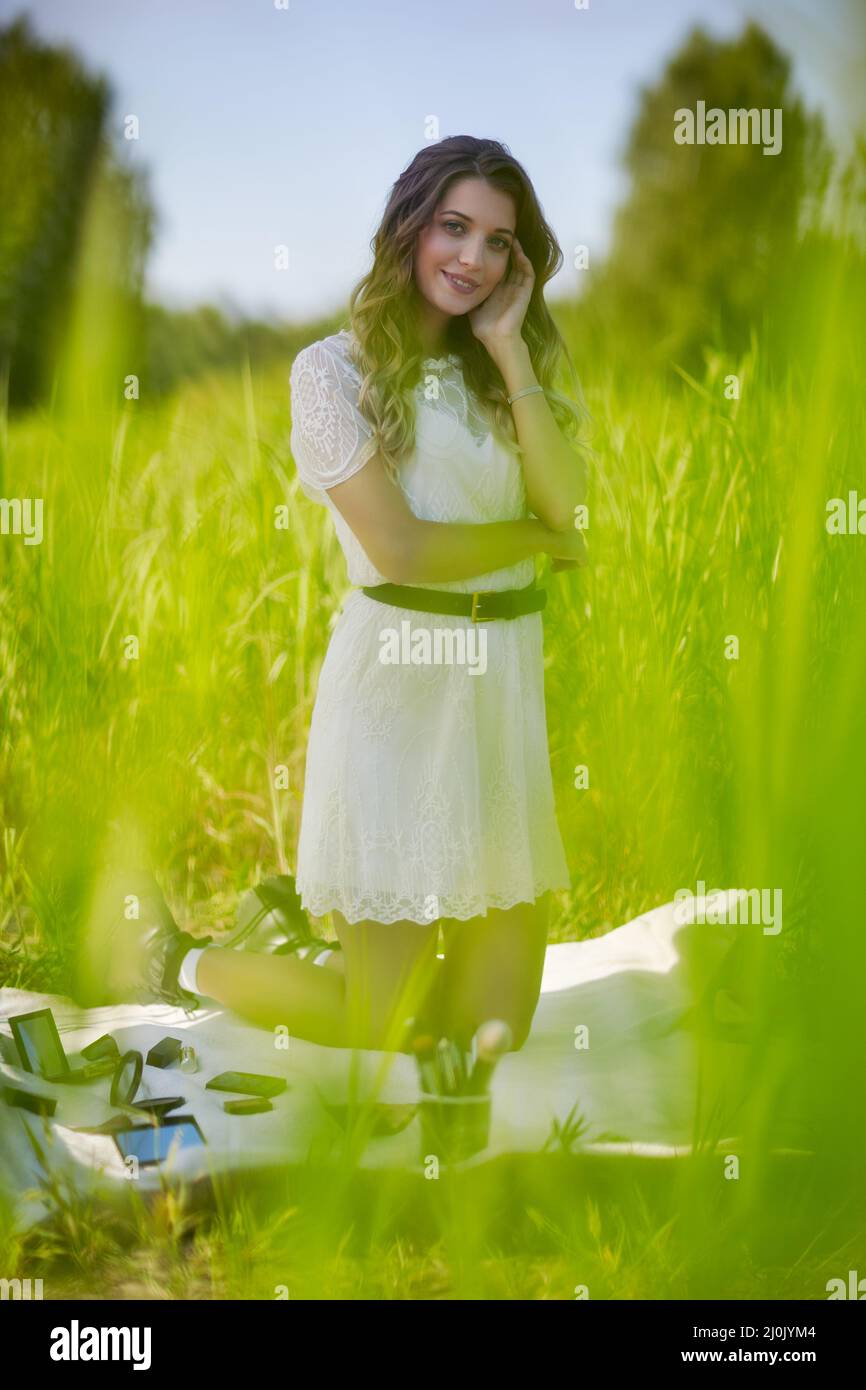 Young blonde woman in white dress is kneeling on a picnic sheet in tall grass. Stock Photo