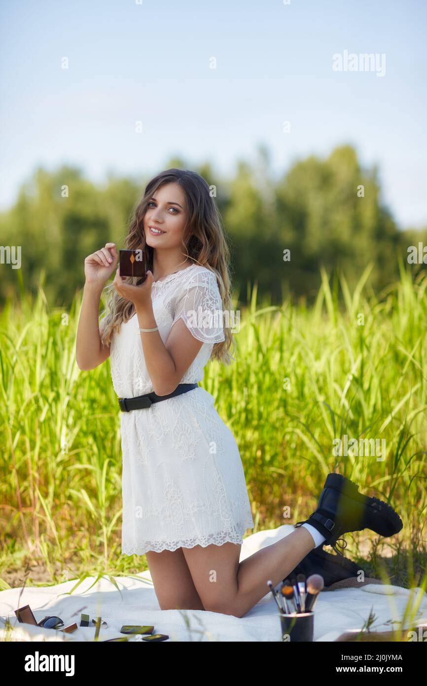 Young blonde woman in white dress is kneeling on a picnic sheet in tall grass. Stock Photo