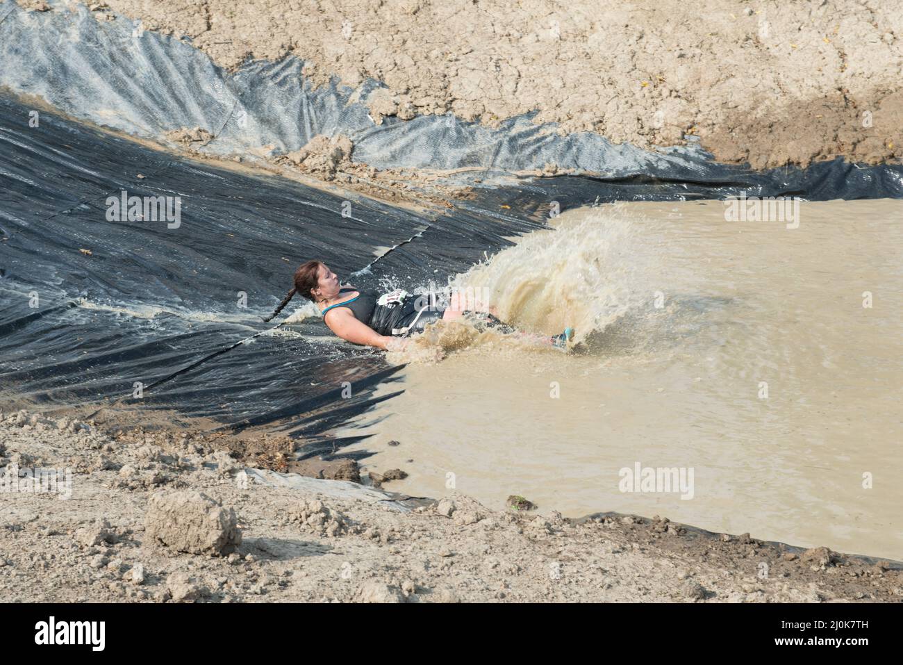 A woman contestant in the4 Swampfoot Run slides into the mud pit feet first with a big splash. Stock Photo