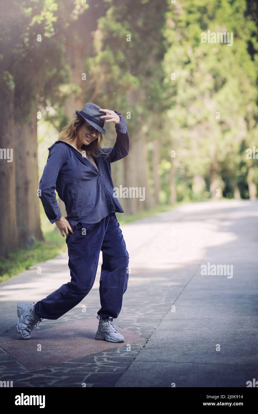 Smiling girl in blue clothes and a hat in a dancing pose in a park alley Stock Photo