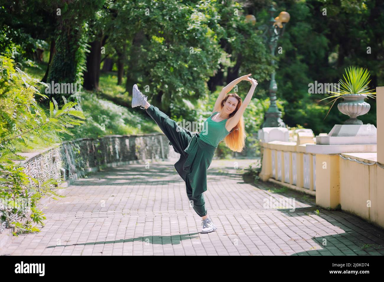 Smiling in a pose with her leg and hands held high in an alley in the park. Dance pose Stock Photo