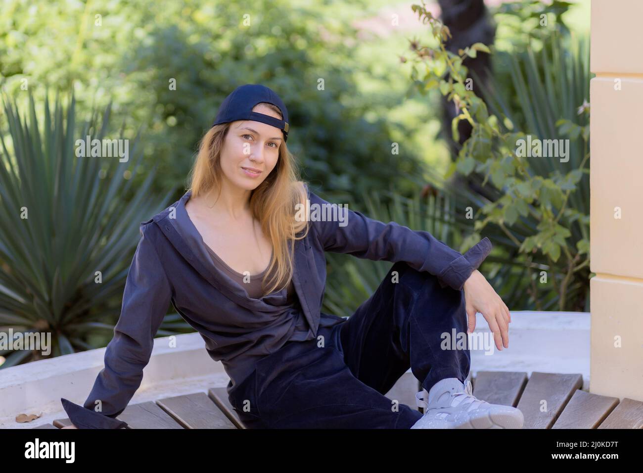 Smiling blonde sports girl sitting on a wooden seat in the park against a background of green southern plants Stock Photo