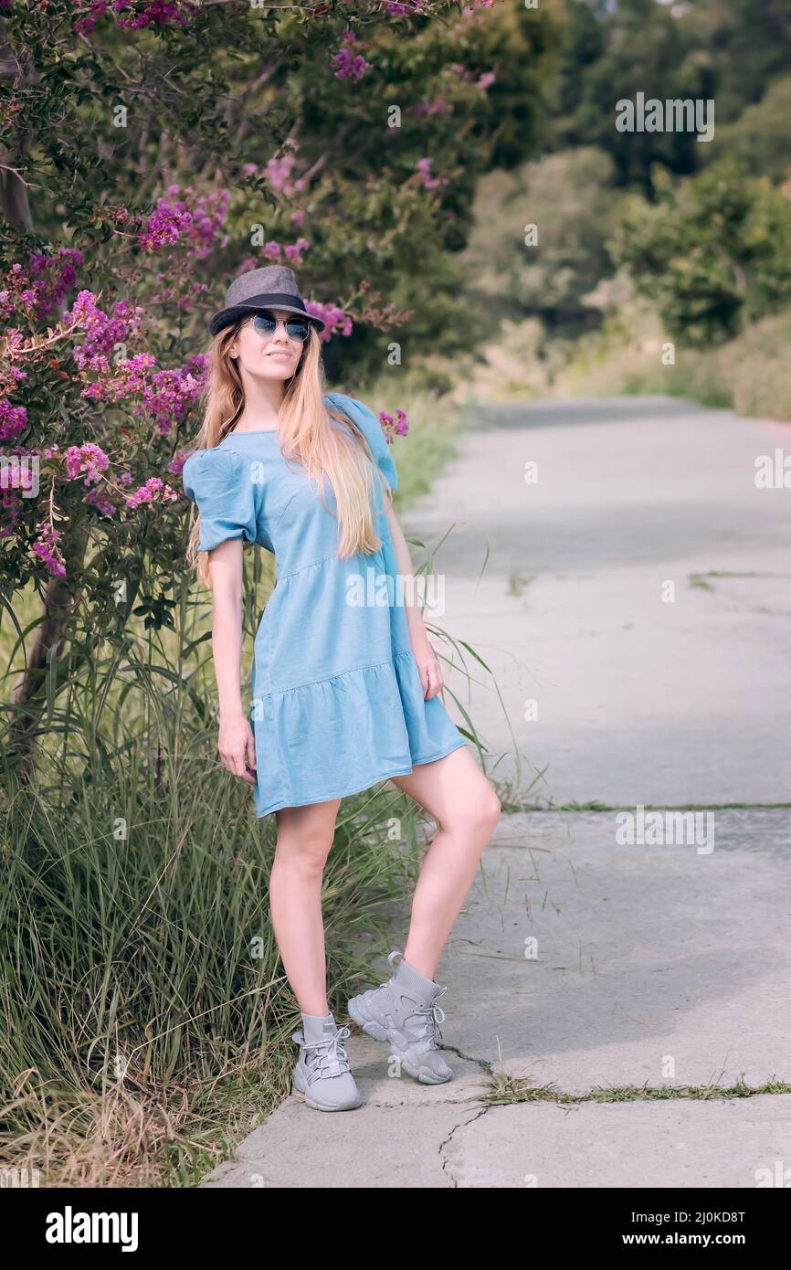 A long-haired girl in a hat and denim dress stands on a concrete alley near a blossoming tree in the park Stock Photo