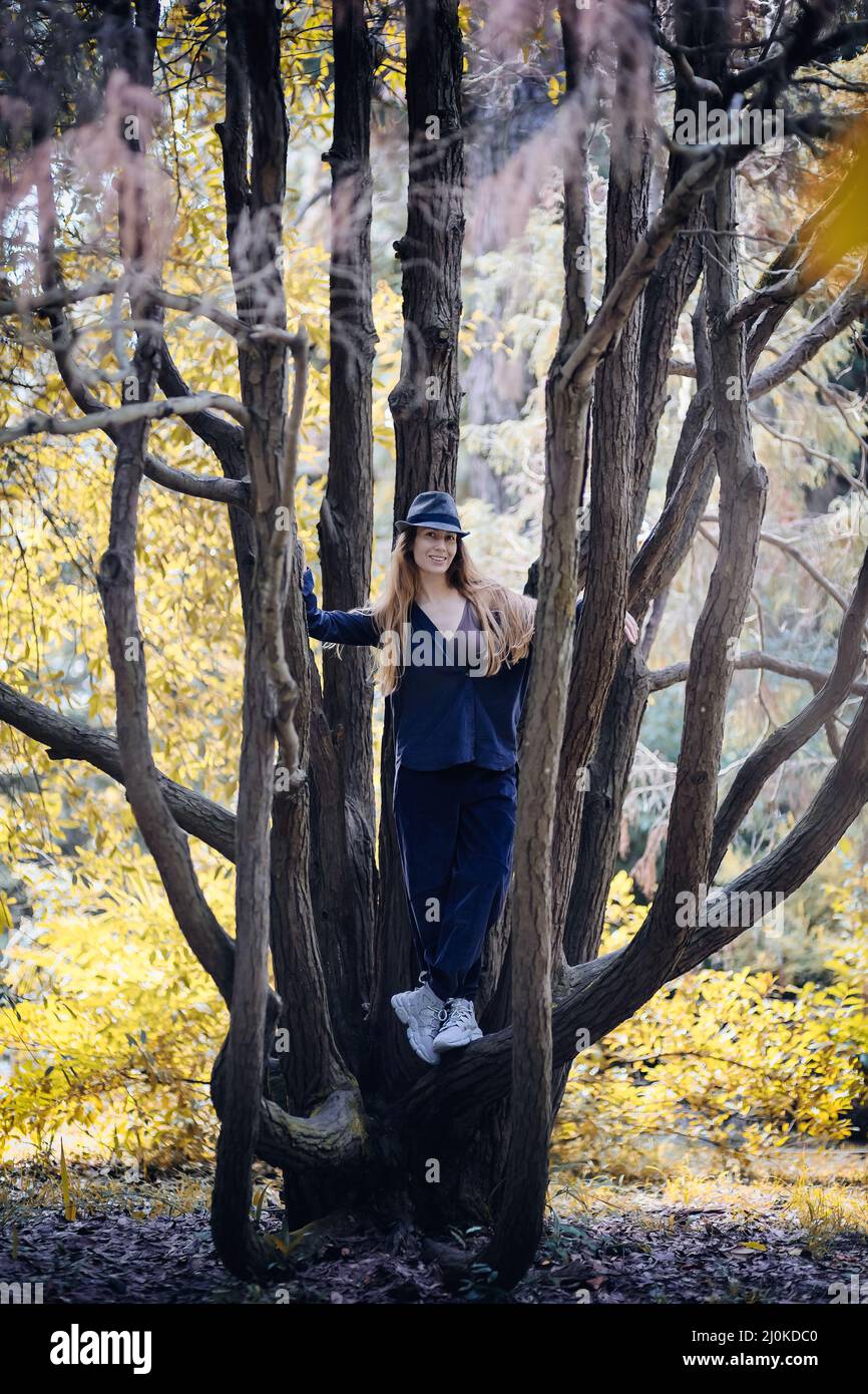Smiling girl in hat and blue casual clothes stands in the center of a branchy tree in an autumn park Stock Photo