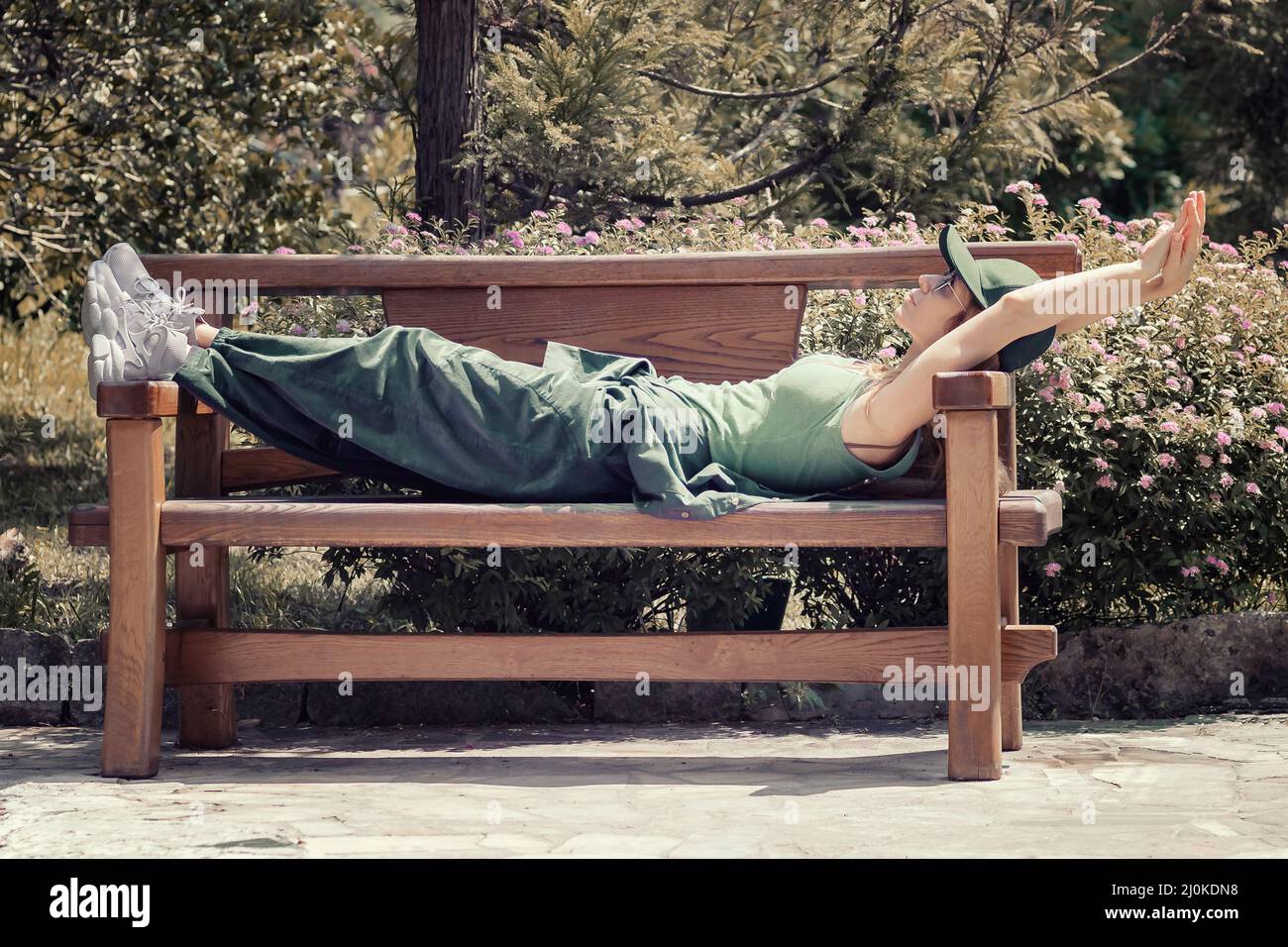 A girl in a green outfit lies on a wooden bench in a summer park Stock Photo