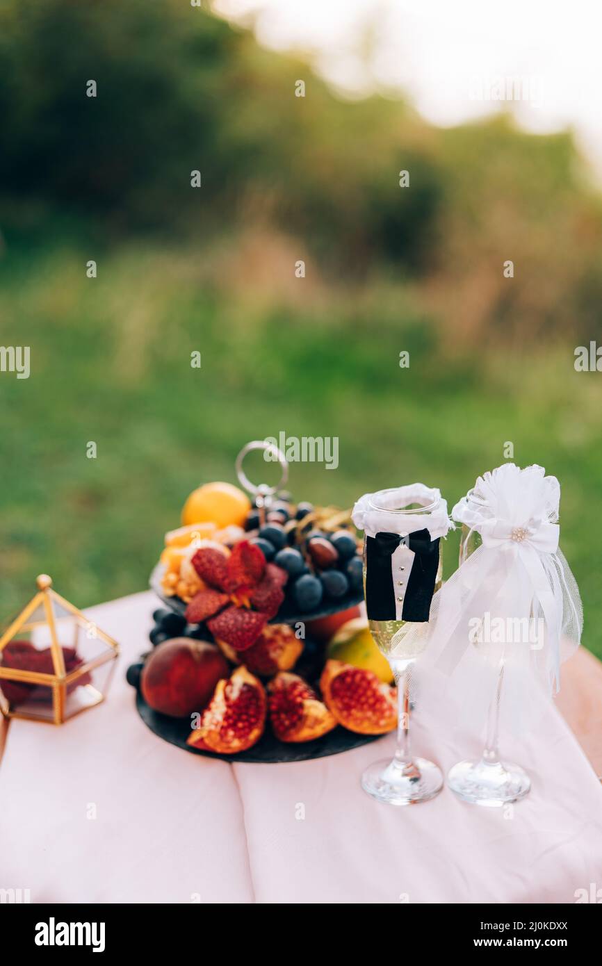 A painted table with a bookcase with fruits and glasses for champagne in the suits of the bride and groom. Stock Photo