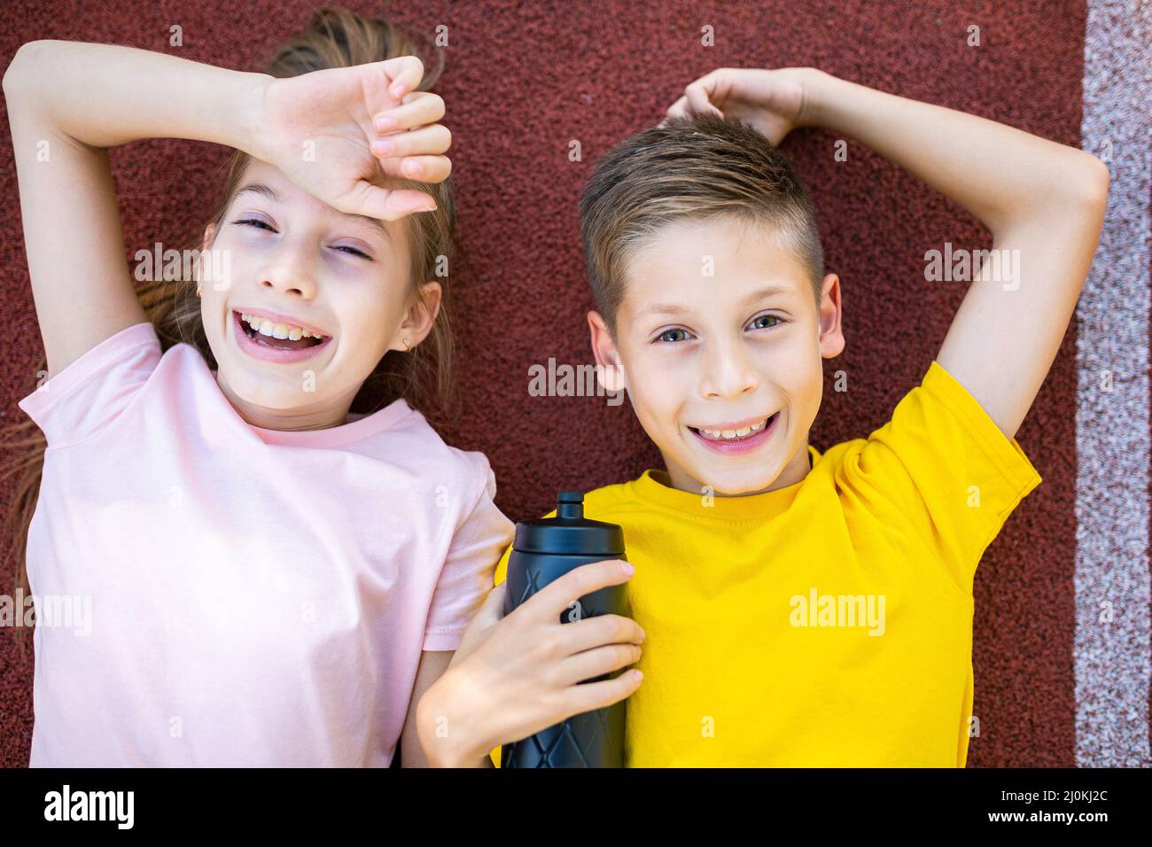 Children sport lifestyle theme. Happy boy and girl teenagers lie joyfully on rubber cover of running track of stadium and erase Stock Photo