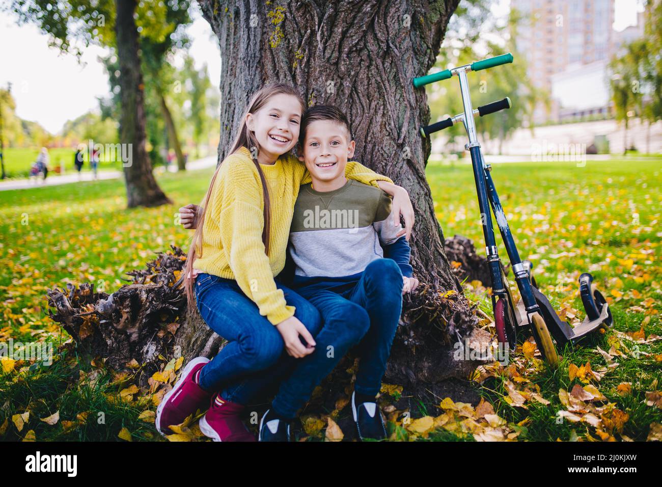 Friends kids posing happily sitting under a tree in an autumn park next to kick scooters. Happy children. Eco transport. Outdoor Stock Photo