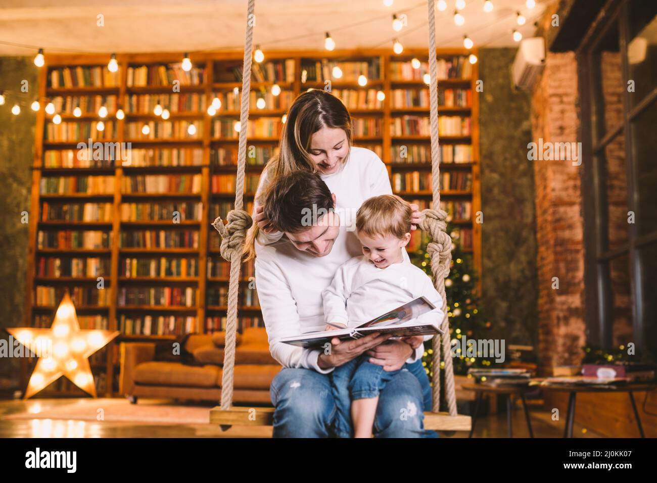 Family With Baby Relaxing On home Swing Seat in cozy dark living room on Christmas eve. Family having fun on seesaw. Winter even Stock Photo