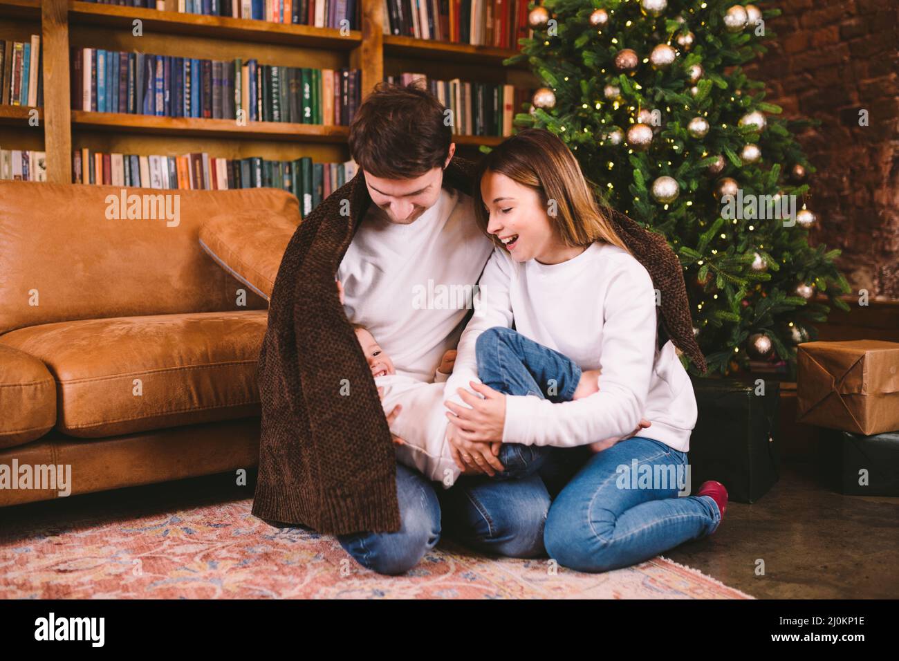 Happy family on Christmas eve on floor under blanket near Christmas tree in evening at home. Family spending Christmas time wrap Stock Photo