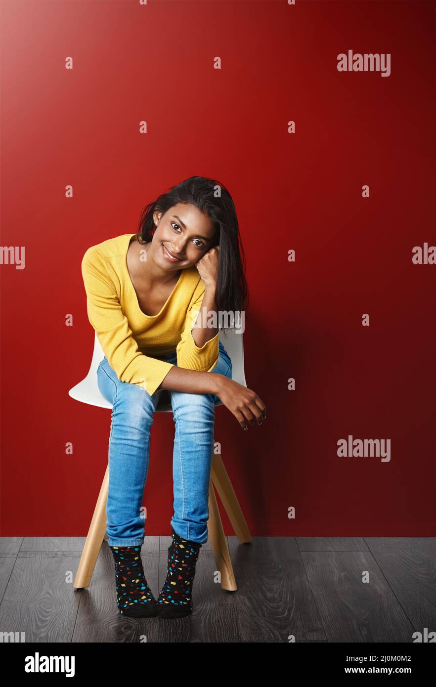 Be just you and youll be beautiful. Studio portrait of a beautiful young woman sitting on a chair against a red background. Stock Photo