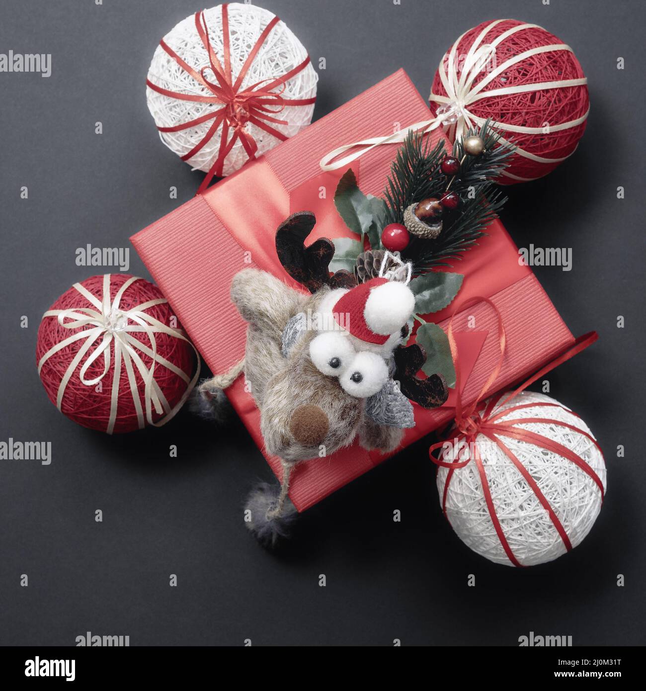 A funny toy big-eyed deer sits on a red gift box with Christmas balls and a coniferous twigs. Christmas composition. Top view Stock Photo