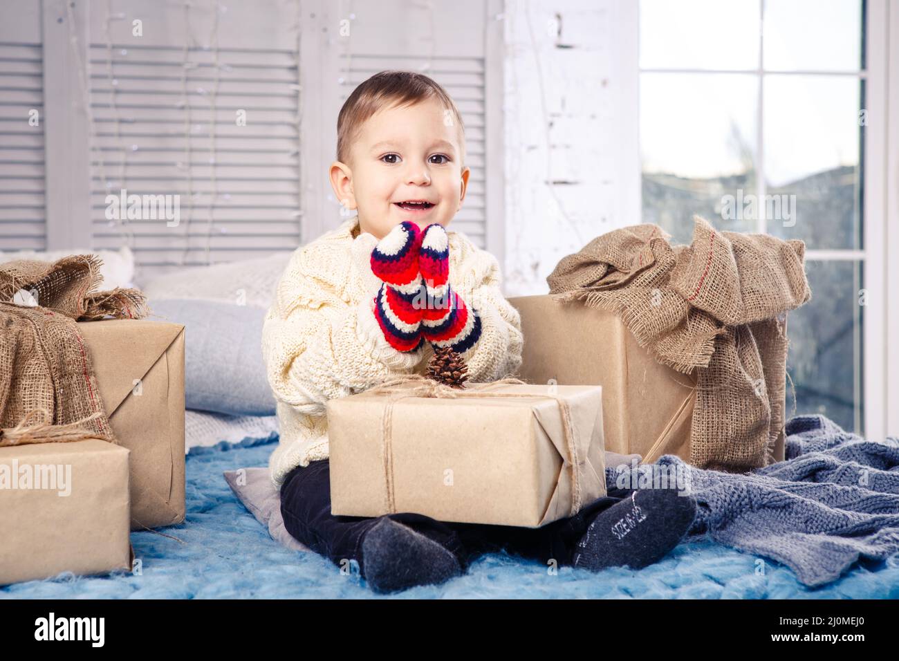 Little funny playful boy a child sits on a bed on Christmas day with gift boxes in white wool knitted sweater and big bright mit Stock Photo