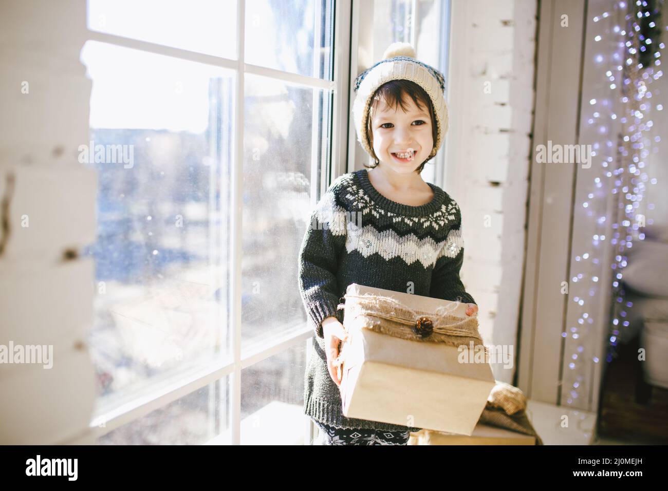 Child is sitting by the window on a sunny Christmas day and makes out with gifts in boxes wrapped in paper.dressed in knitted wa Stock Photo